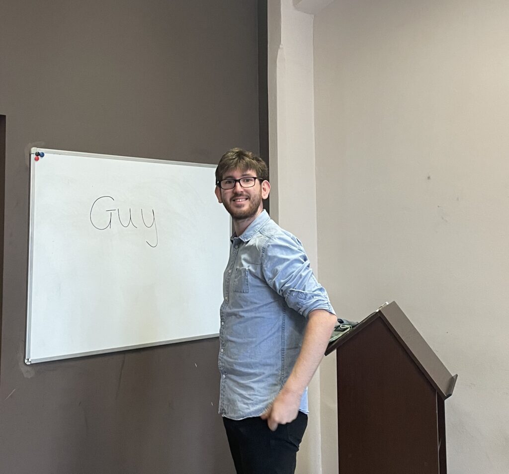 Mężczyzna w koszuli stoi obok tablicy z napisem Guy, uśmiechając się do kamery